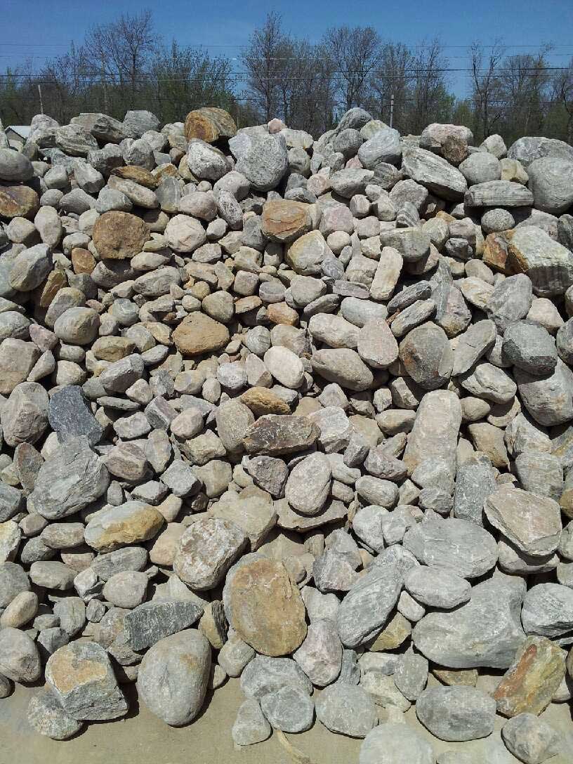 Large pile of gray and tan rocks outdoors with a blue sky above the yard fence