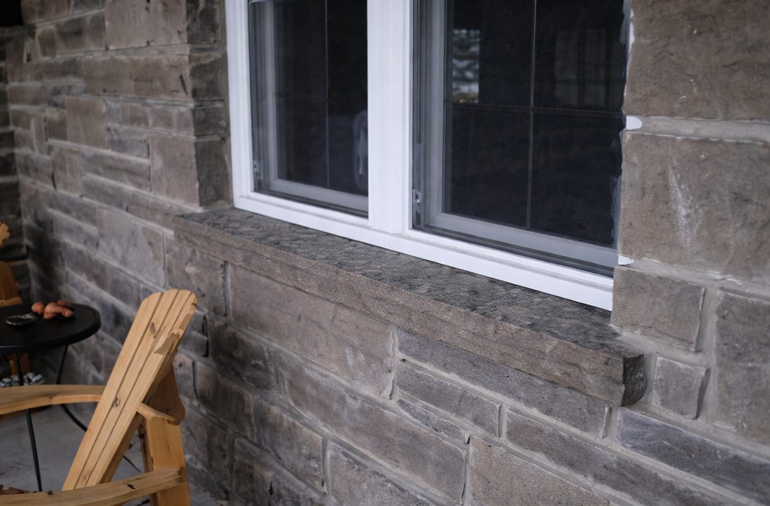 Stone exterior wall with a white-framed window and a wooden Adirondack chair on a small patio table nearby.